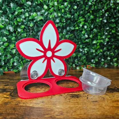 Red and white flower-shaped feeding ledge on a wooden surface with a green leafy background